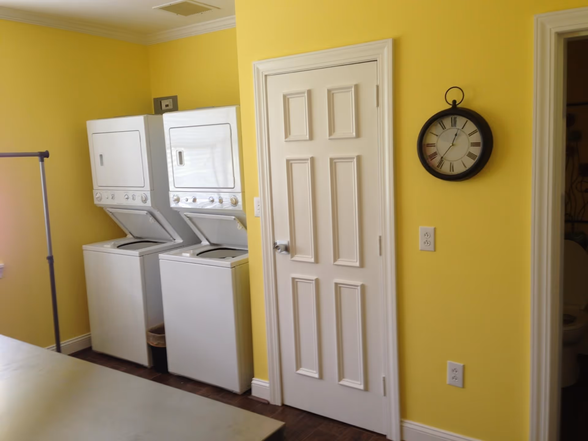 Laundry room with two stacked white washer and dryer units against a yellow wall. There is a white door in the center and a round wall clock with Roman numerals hanging on the wall to the right. A small part of a toilet is visible through an open doorway on the far right.