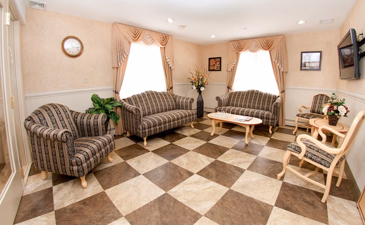 A cozy living room with patterned armchairs and sofas arranged around a wooden coffee table. The room has checkered brown and beige flooring, two large windows with beige curtains, a wall clock, framed pictures, a flat-screen TV mounted on the wall, and decorative plants and flowers.