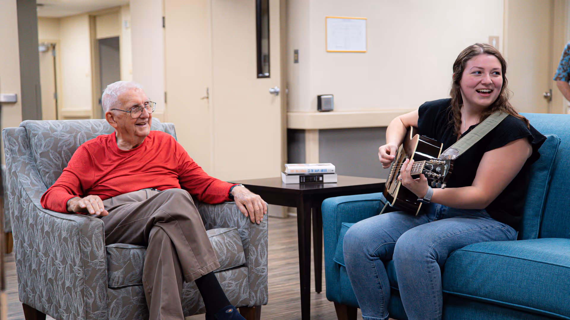An elderly man sits in an armchair smiling while a young woman plays an acoustic guitar on a nearby couch in a common room.