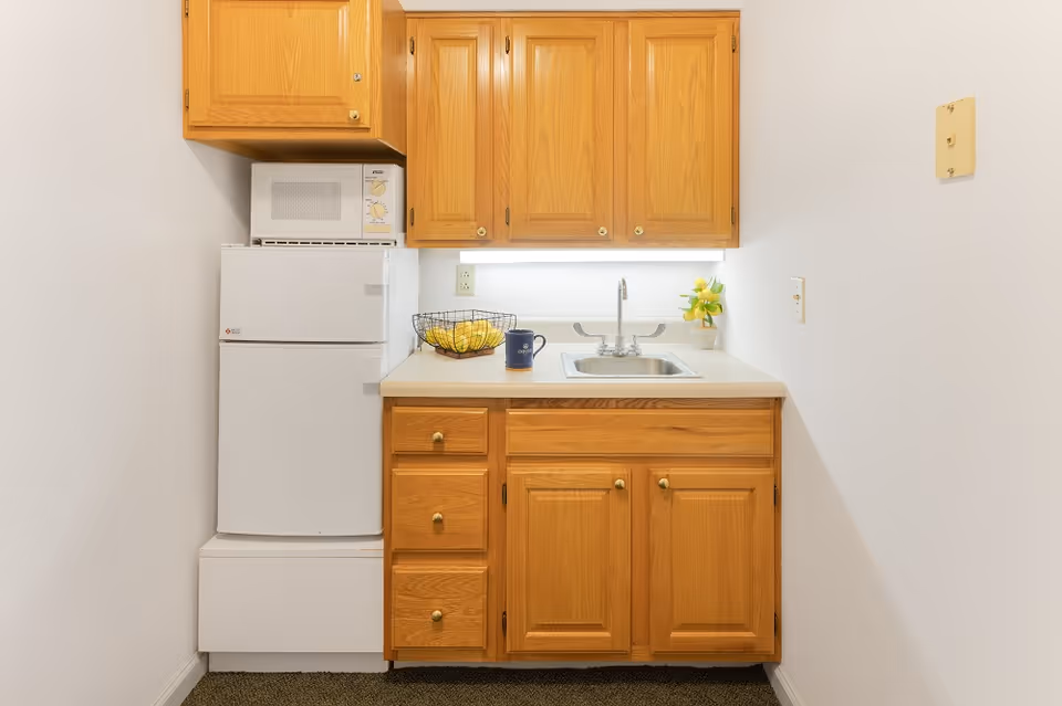 Small kitchenette with wooden cabinets, a white mini refrigerator, a microwave on top of the refrigerator, a sink with a faucet, a wire basket with lemons, a blue mug, and a small plant with yellow flowers on the countertop.