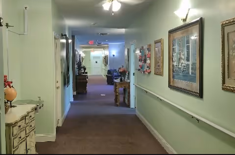 A long, carpeted hallway in an assisted living facility with light green walls, handrails on both sides, framed artwork hanging on the right wall, and a ceiling fan with lights. There are tables with decorative items along the hallway and doors leading to rooms on the left side.