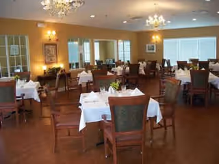A spacious dining room with multiple tables covered in white tablecloths, each set with folded napkins and small flower arrangements. The room has wooden chairs with green upholstery, large windows letting in natural light, chandeliers, and framed artwork on the walls.