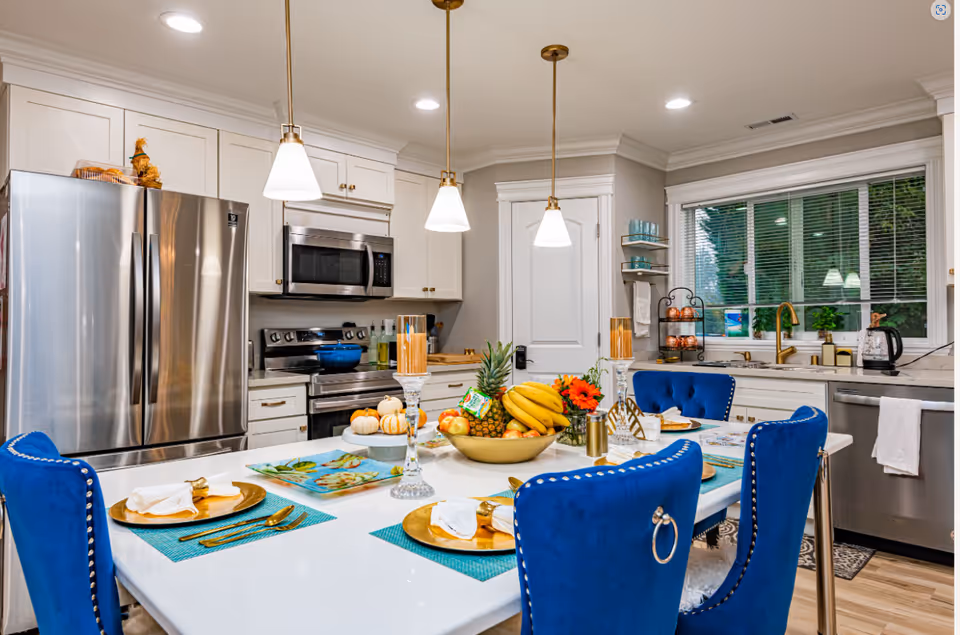 A modern kitchen with white cabinets, stainless steel refrigerator, stove, microwave, and dishwasher. A white countertop island with blue upholstered chairs is set with gold plates, white napkins, and gold cutlery. The island also features a centerpiece bowl with bananas, a pineapple, and other fruits, along with two tall candle holders and a small flower arrangement. Three pendant lights hang above the island, and a window with blinds is visible above the sink.