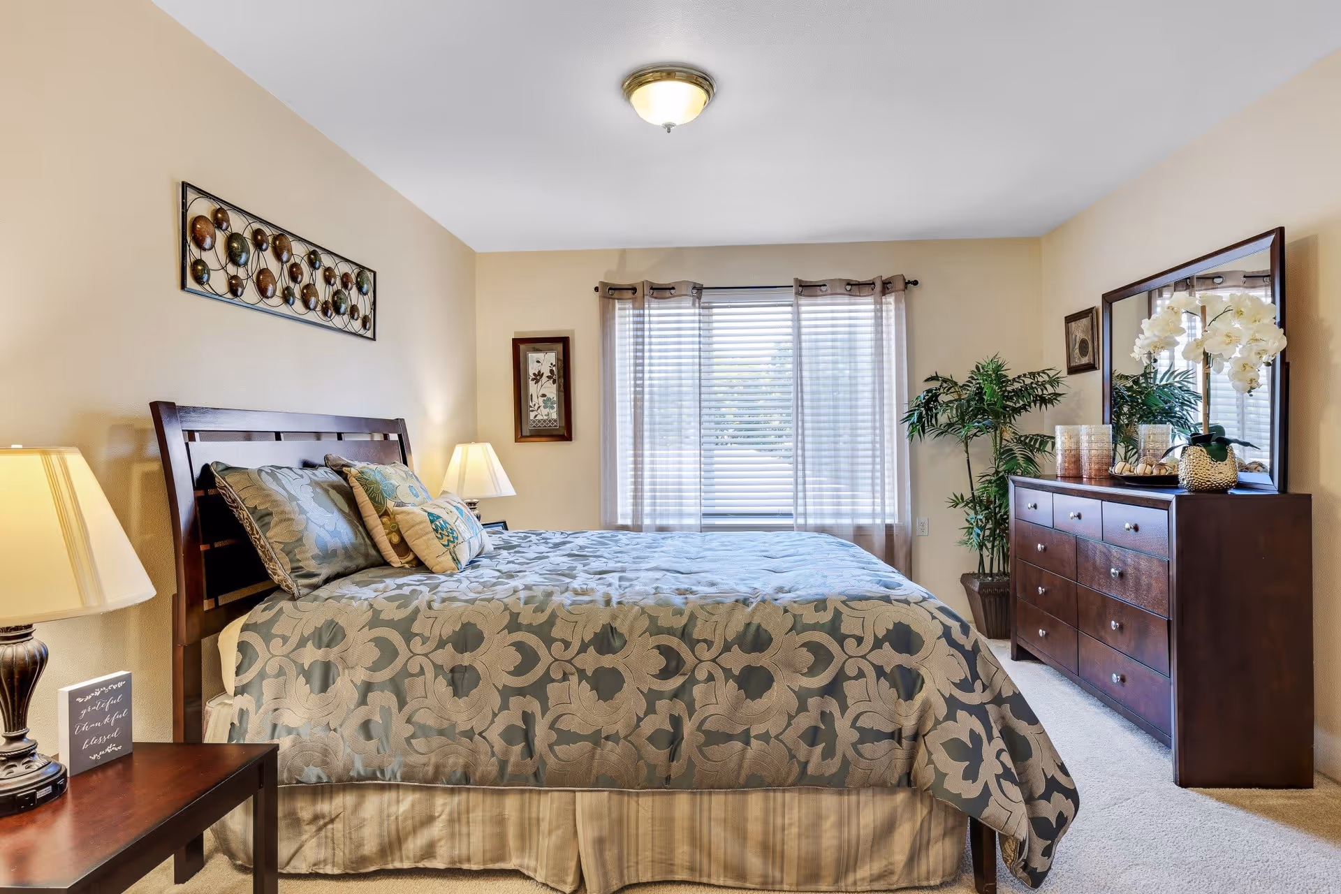 A neatly made bed with patterned bedding in a bedroom featuring beige walls, a wooden headboard, two bedside tables with lamps, a large window with sheer curtains, a wooden dresser with a mirror, decorative plants, and wall art.