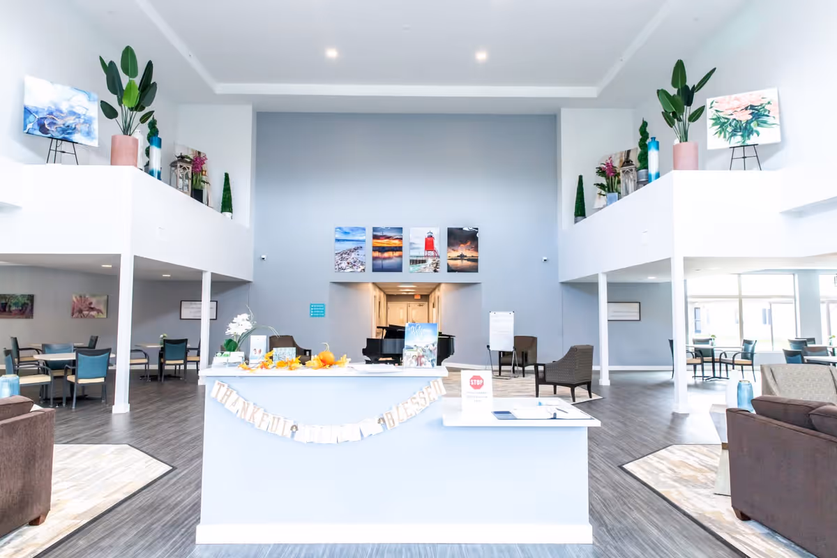 Bright and spacious senior living facility common area with a reception desk decorated with a 'Thankful, Grateful, Blessed' banner. The room features high ceilings, modern furniture including chairs and tables, plants, and colorful artwork on the walls. There is a piano in the background and large windows allowing natural light to fill the space.