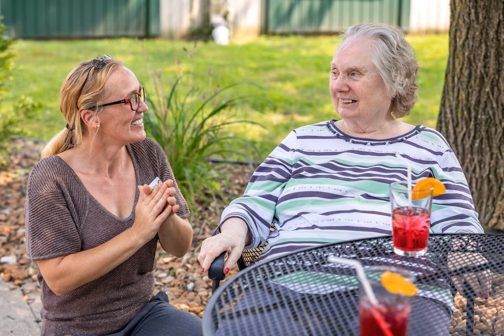 An elderly woman sitting at an outdoor metal table with two red drinks garnished with orange slices, smiling and looking at a woman with glasses and a ponytail who is kneeling beside her and smiling back. They are in a garden area with green grass and plants in the background.