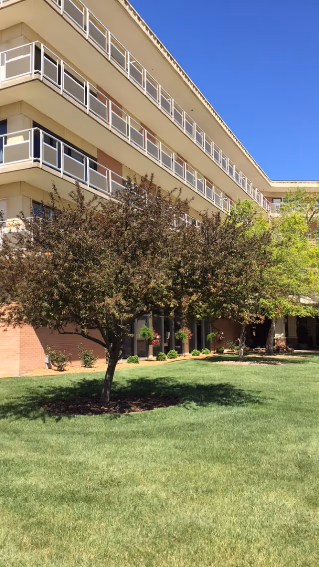 Exterior view of a multi-story senior living facility building with balconies, a well-maintained green lawn, and several trees in the foreground under a clear blue sky.