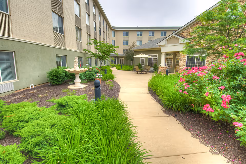Outdoor courtyard area at Westside Garden Plaza featuring a paved walkway surrounded by green shrubs, flowering plants, and a decorative fountain. The building exterior has multiple windows and a seating area with a table and umbrella near the entrance.