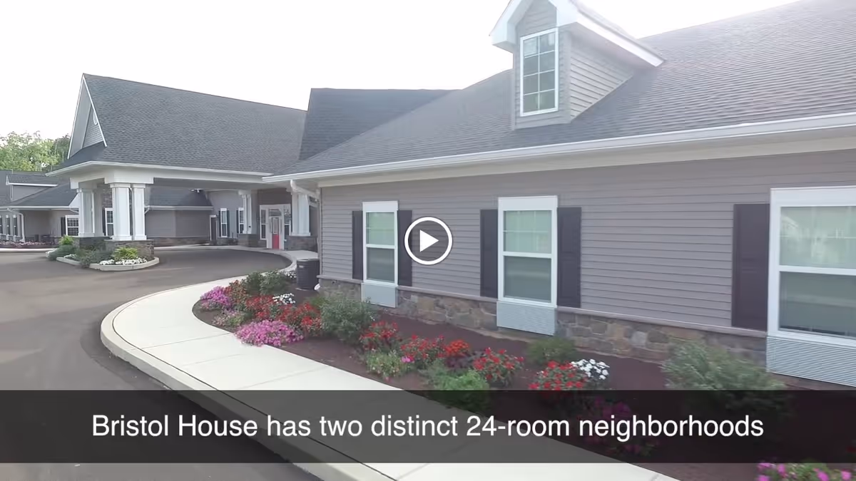Exterior view of a single-story building with gray siding, white trim, and a dark roof. There is a curved driveway and a sidewalk bordered by colorful flower beds in front of the building. The building has multiple windows with black shutters and a small dormer window on the roof.