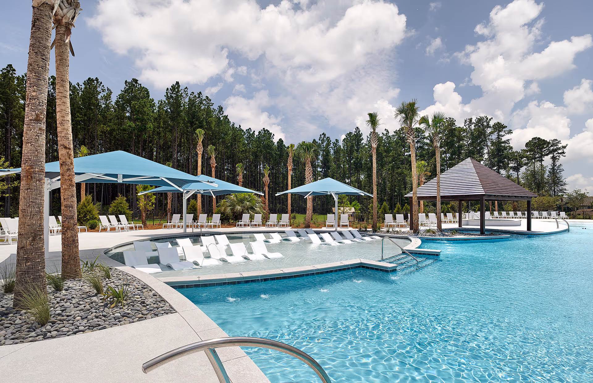 Outdoor swimming pool area with lounge chairs partially submerged in shallow water, large blue umbrellas providing shade, palm trees, and a covered pavilion. The pool is surrounded by a concrete deck and lush greenery with tall trees in the background under a partly cloudy sky.