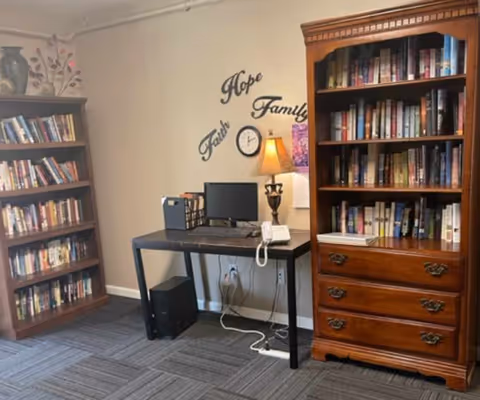 A cozy interior room with two wooden bookshelves filled with books on either side of a black desk. The desk holds a computer monitor, a lamp, a telephone, and some office supplies. On the wall above the desk are decorative words 'Faith', 'Hope', and 'Family' along with a small clock. The floor is carpeted with a patterned design.