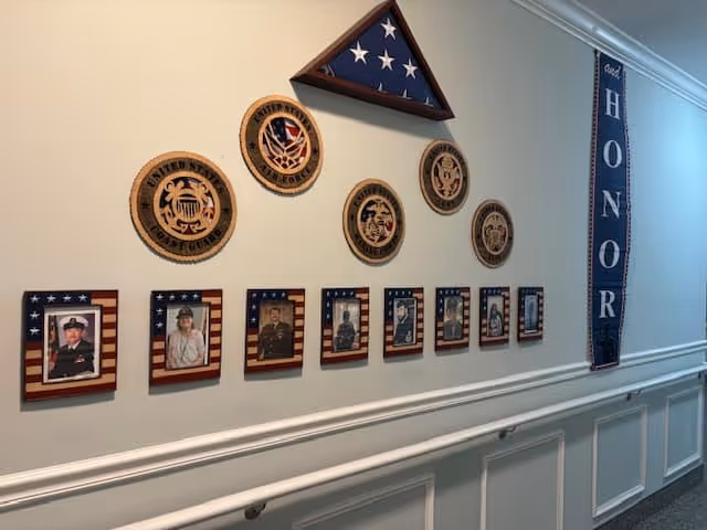 Hallway wall display with a folded American flag, military seals, framed veteran photos, and a vertical 'HONOR' banner.