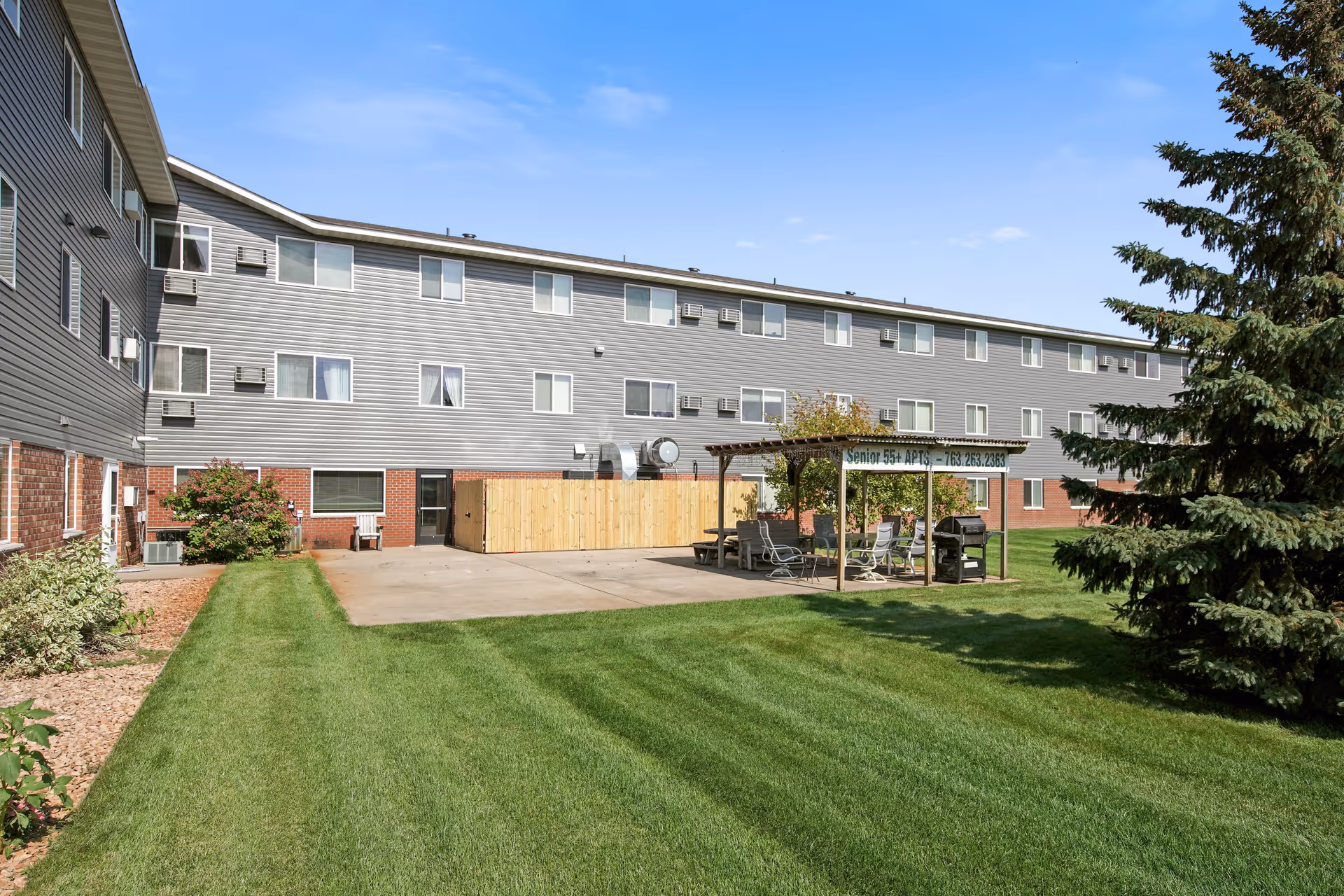 Outdoor view of a senior living facility named Keller Lake Commons showing a three-story building with multiple windows and air conditioning units. In front of the building is a well-maintained grassy area with a covered patio that has chairs, tables, and a grill. A large evergreen tree is visible on the right side under a clear blue sky.