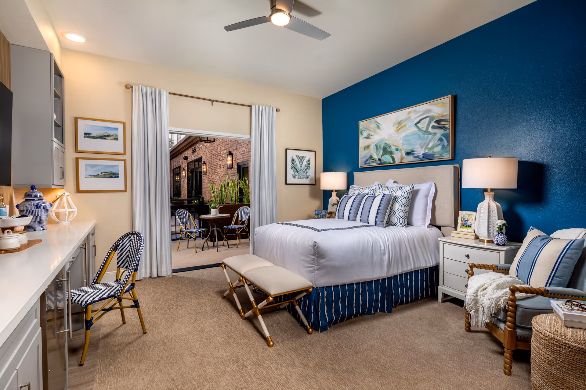 A cozy bedroom with a bed featuring white and blue bedding, a beige bench at the foot of the bed, a nightstand with a lamp and framed photo, a comfortable armchair with a striped pillow and throw blanket, and a desk area with a chair. The room has a blue accent wall with abstract artwork and an open sliding door leading to an outdoor patio with a small table and two chairs.