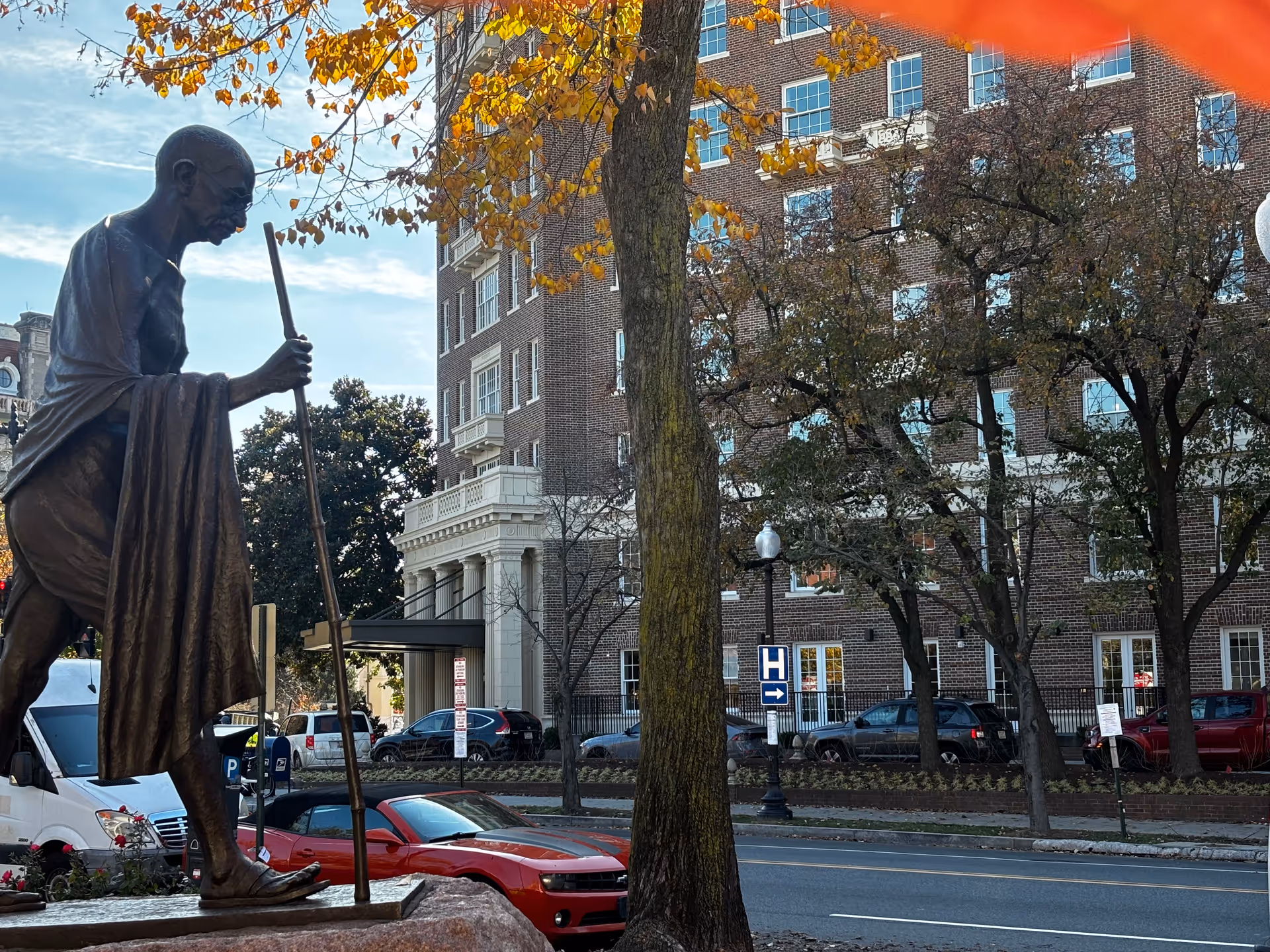 A bronze statue of Mahatma Gandhi holding a walking stick, positioned on a stone pedestal near a street. Behind the statue, there is a large brick building with white window frames and architectural details. Trees with autumn-colored leaves partially obscure the building. Several parked cars line the street, and a street sign with a hospital symbol and an arrow is visible.