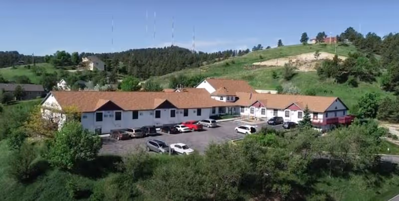 Aerial view of a single-story assisted living facility with a red roof surrounded by greenery and hills. Several cars are parked in the parking lot in front of the building.