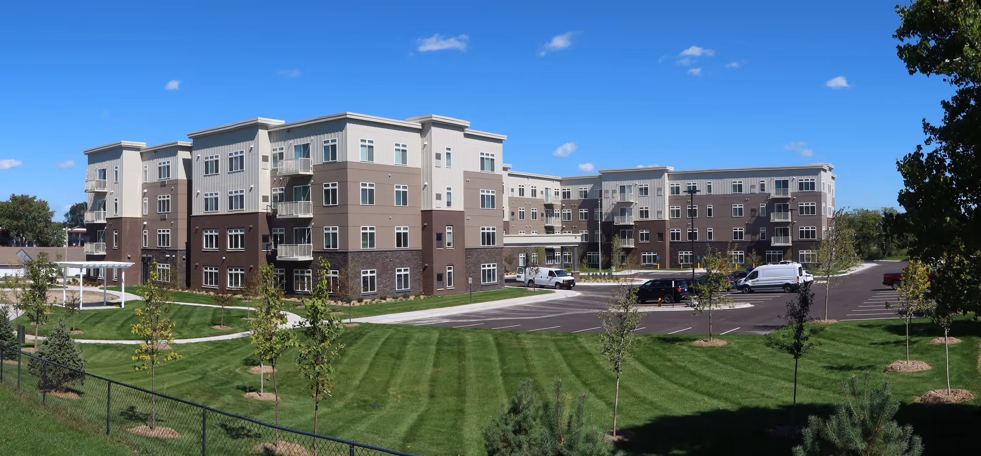 Exterior view of a modern four-story senior living facility building with a well-maintained lawn, young trees, a parking lot with several vehicles, and a clear blue sky.
