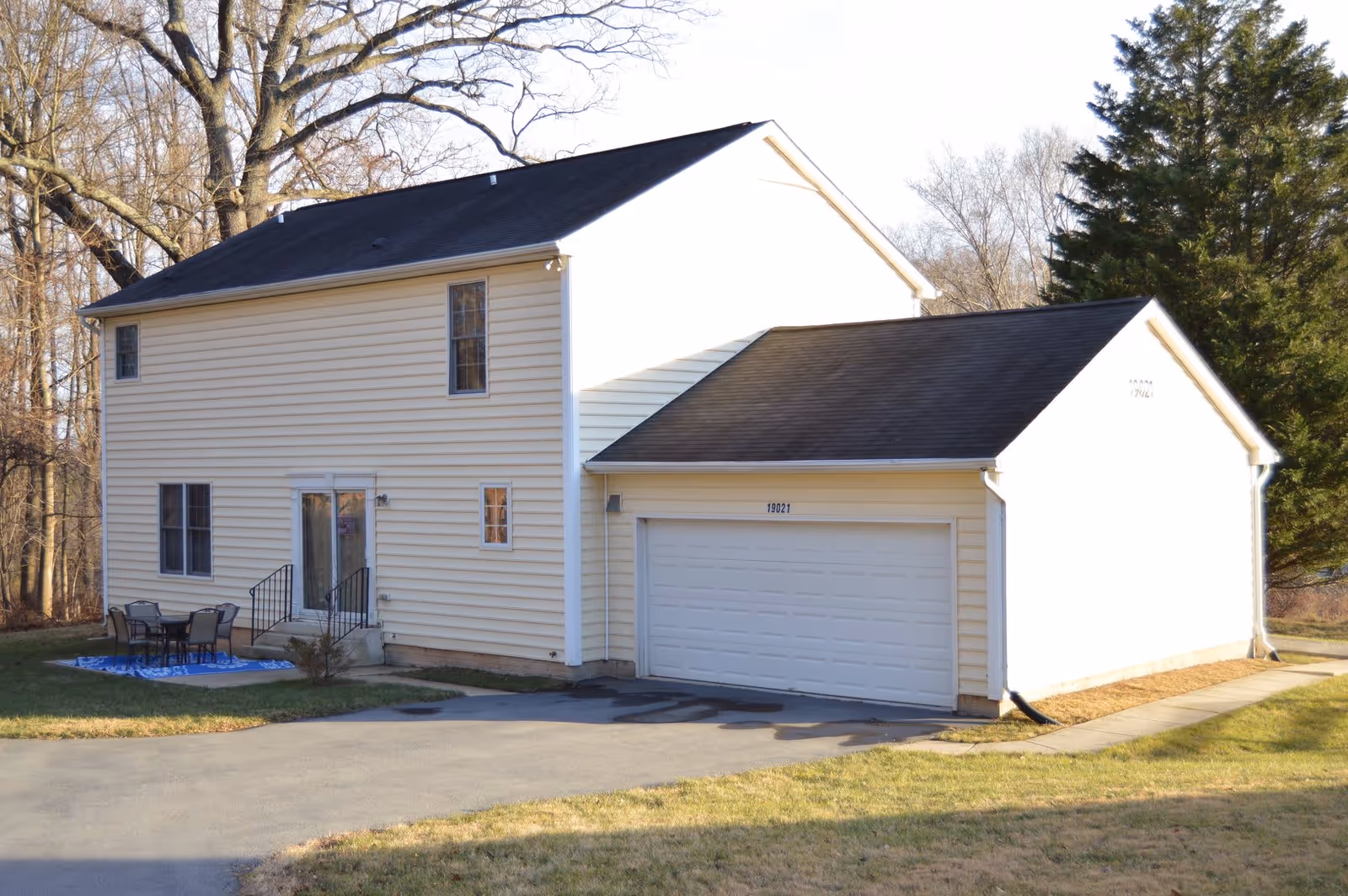 Exterior view of a two-story beige house with a black roof and attached two-car garage. There is a small patio area with a table and four chairs on a blue outdoor rug. Trees without leaves are visible in the background.