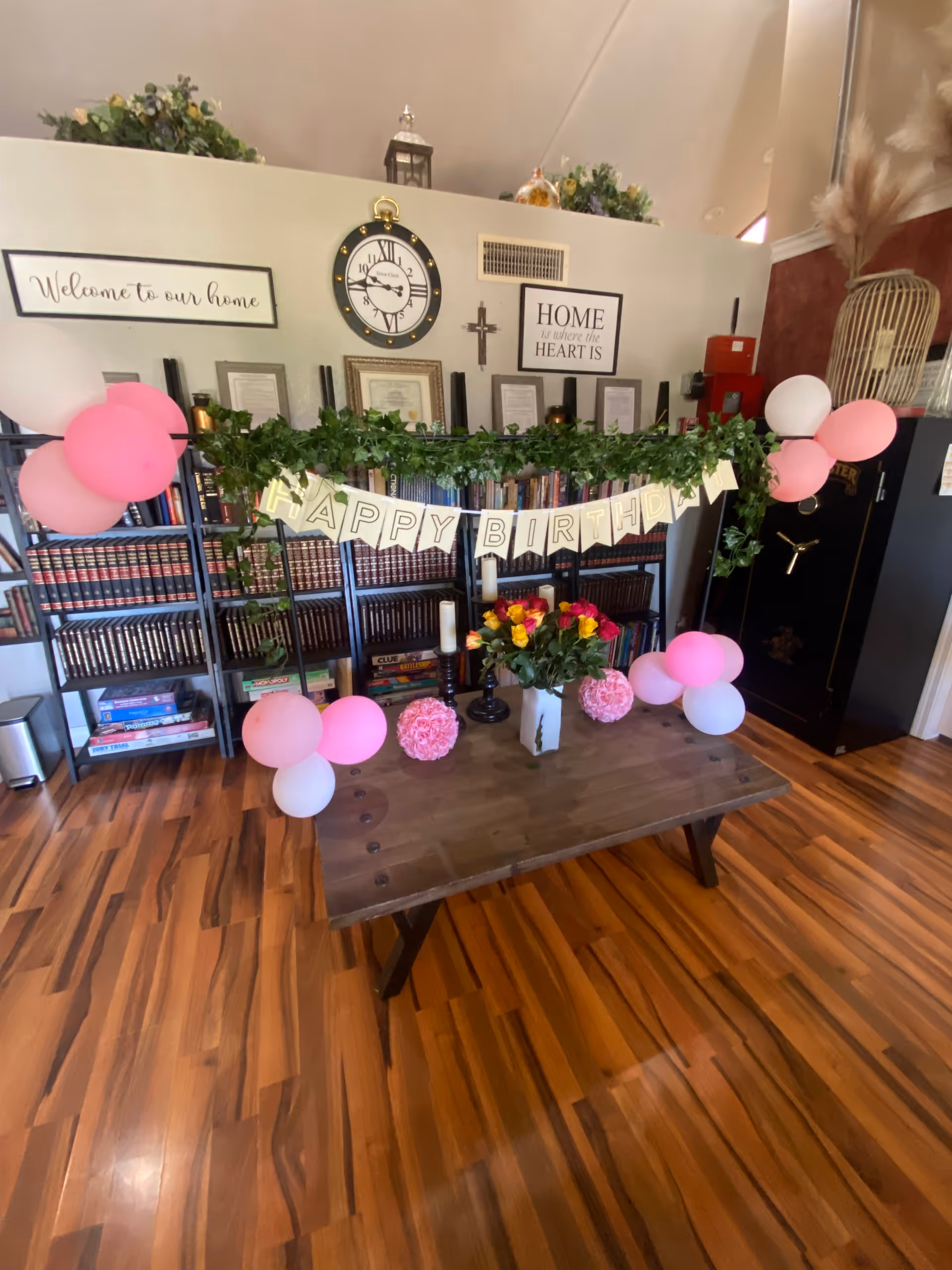 Interior common room with bookshelves and a decorated table displaying pink balloons, a "Happy Birthday" banner, and a vase of flowers.