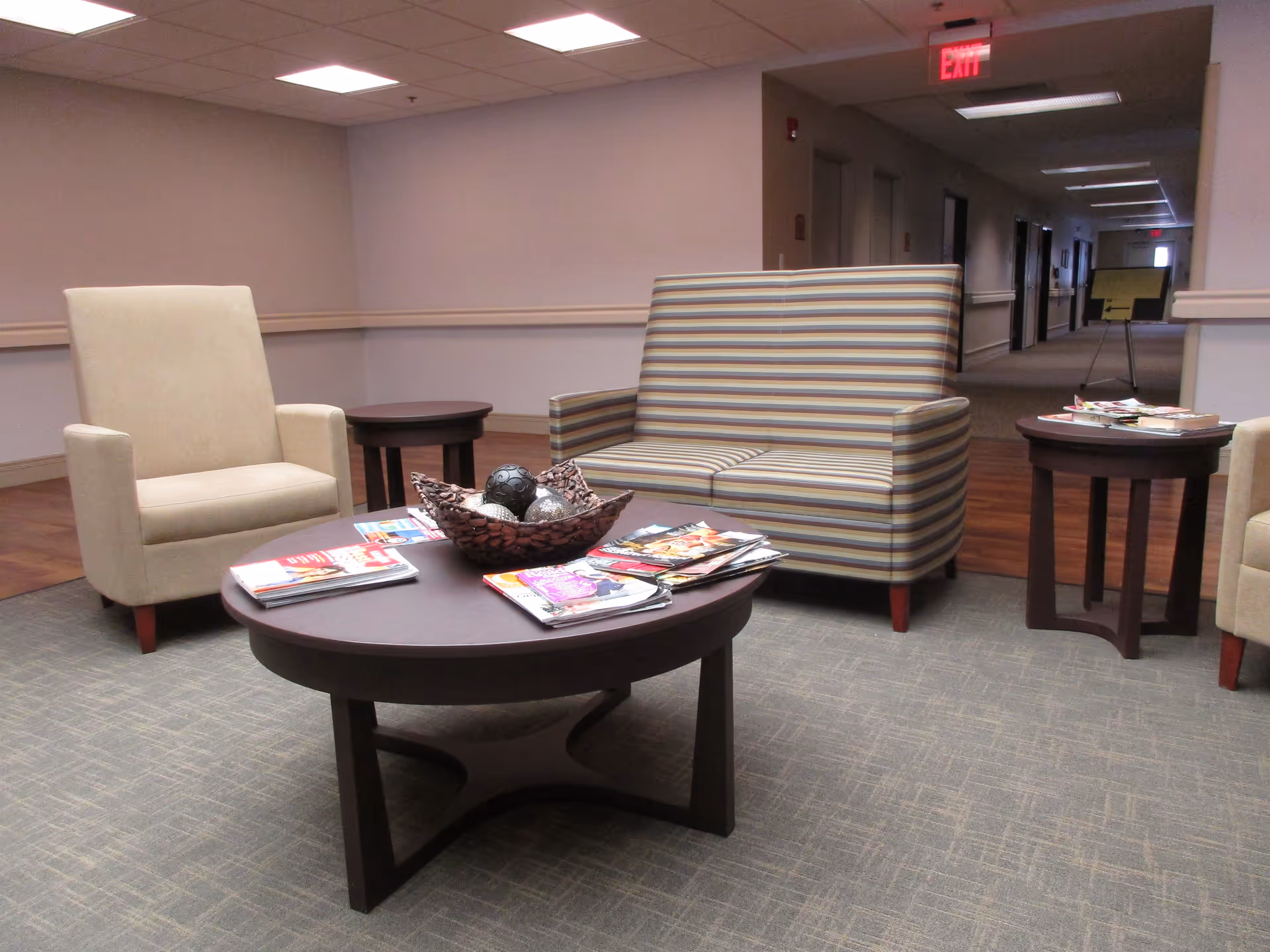 A seating area in a senior living facility with a beige armchair, a striped loveseat, and three dark wooden tables. The tables hold magazines and a decorative bowl with ornamental balls. The area is carpeted and adjacent to a hallway with overhead lighting and an exit sign.