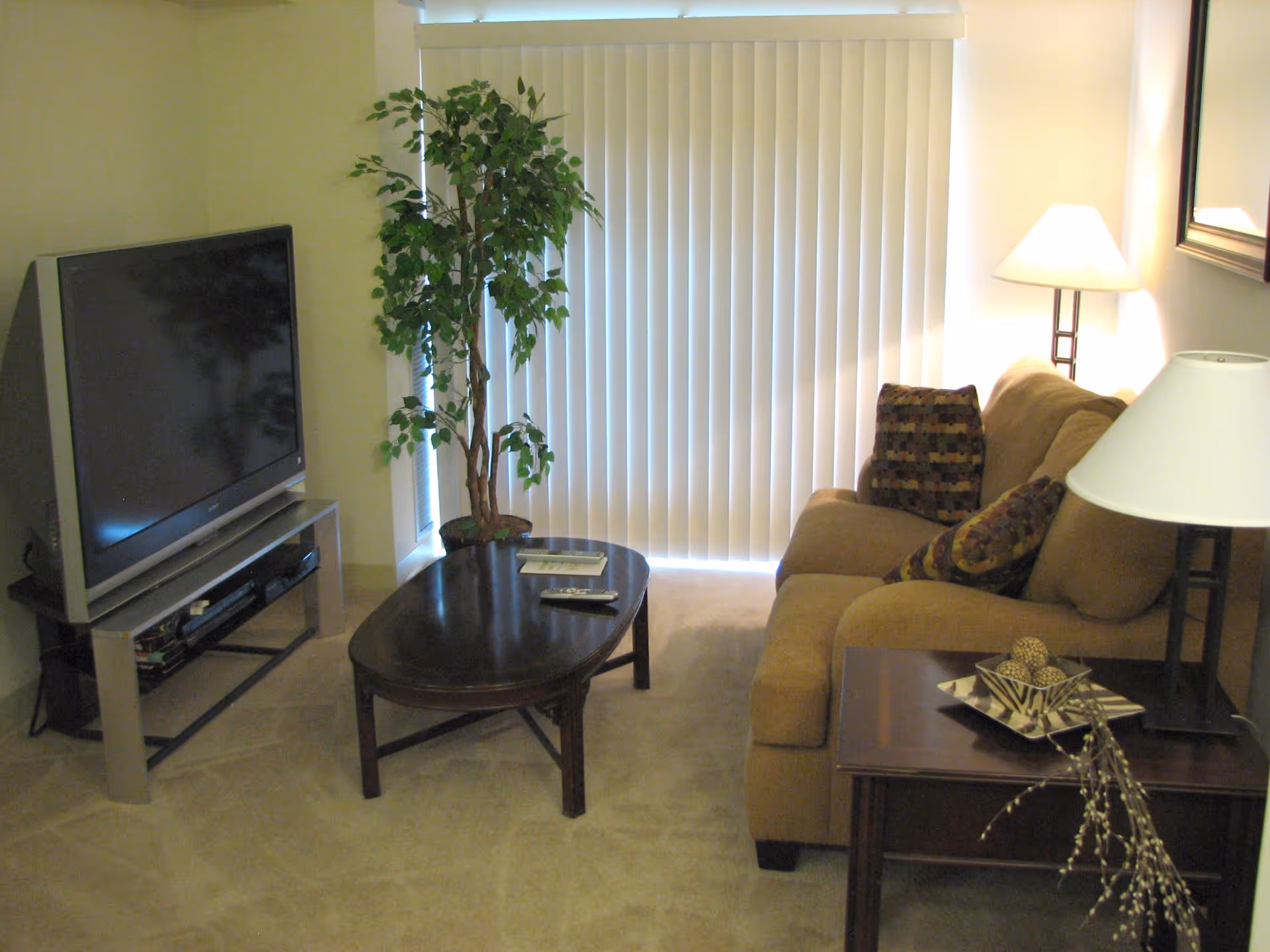A cozy living room with a beige sofa adorned with patterned cushions, a dark wooden coffee table with a remote and a book on it, a tall green potted plant near vertical blinds covering a window, a flat-screen TV on a stand, and two lamps providing warm lighting.