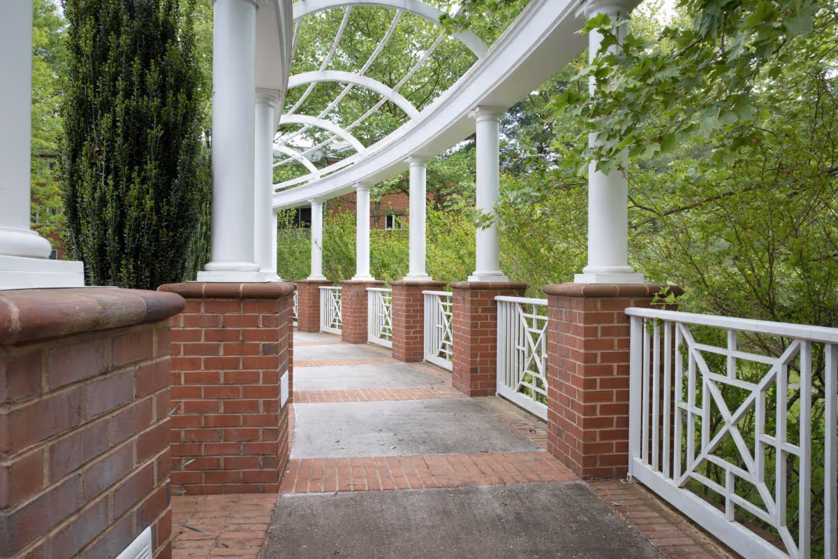 A covered outdoor walkway with white columns and a curved white trellis overhead, bordered by red brick pillars and white railings, surrounded by green trees and shrubs.