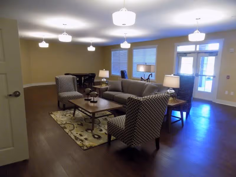 A well-lit living room area with a gray sofa, two patterned armchairs, a wooden coffee table on a patterned rug, two side tables with lamps, and a dining table with chairs in the background. The room has hardwood floors, beige walls, multiple ceiling lights, and a glass door with windows letting in natural light.