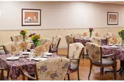 A dining room with several tables covered in purple patterned tablecloths, each set with plates, cups, and napkins folded in a fan shape. The chairs have floral upholstery, and there are small flower arrangements on each table. Two framed pictures hang on the beige walls.