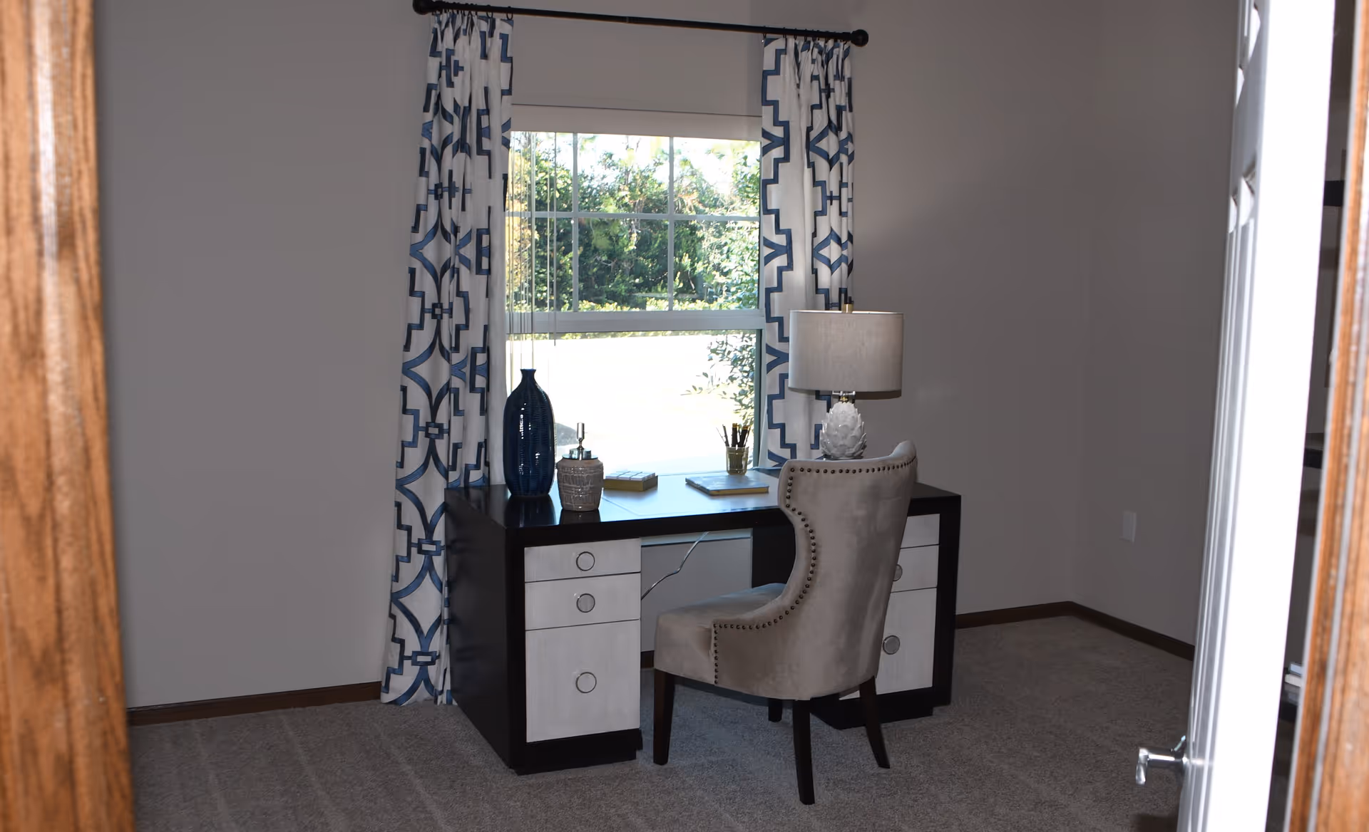 A small home office setup with a black and white desk placed in front of a window with blue and white patterned curtains. On the desk are decorative items including a blue vase, a small container, books, and a lamp with a white shade. A beige upholstered chair with nailhead trim is positioned in front of the desk. The room has beige carpet and light-colored walls.