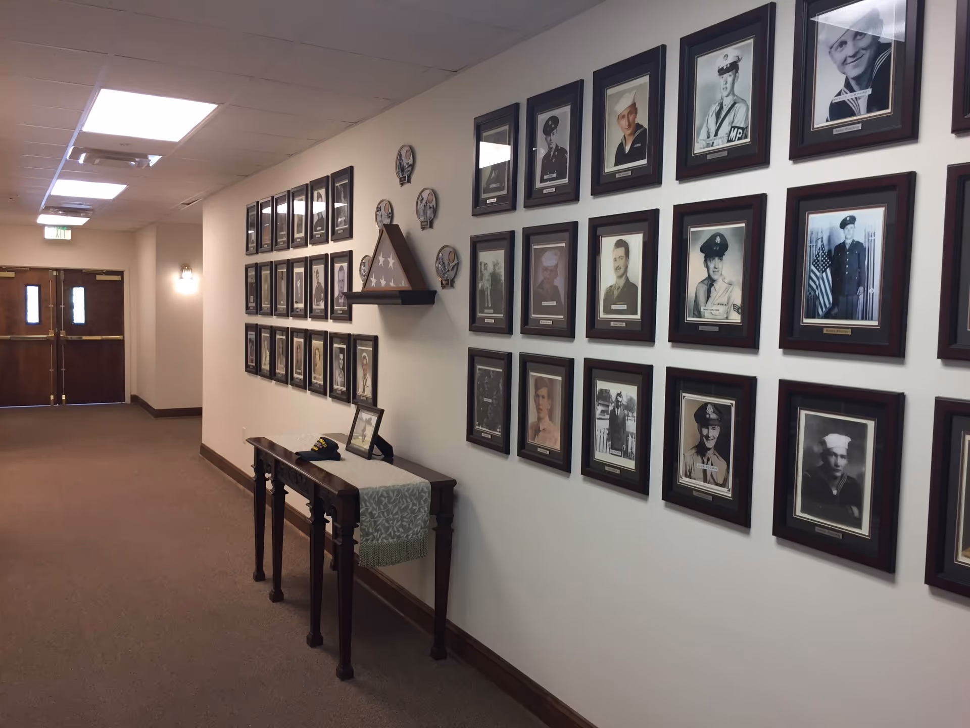A hallway in a senior living facility with framed black-and-white portraits of military veterans displayed on the wall. Below the portraits is a wooden table with a decorative runner, a folded American flag in a triangular display case, a framed photo, and a military cap. The hallway has carpeted floors, beige walls, and double wooden doors at the end.