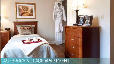 A cozy bedroom in Ashbrook Village featuring a single bed with a beige blanket and decorative pillows. A tray with a cup and napkin is placed on the bed. On the wall above the bed is a framed landscape painting. To the right, there is a wooden dresser with a lamp and framed photos on top. A white robe hangs on the door in the background.