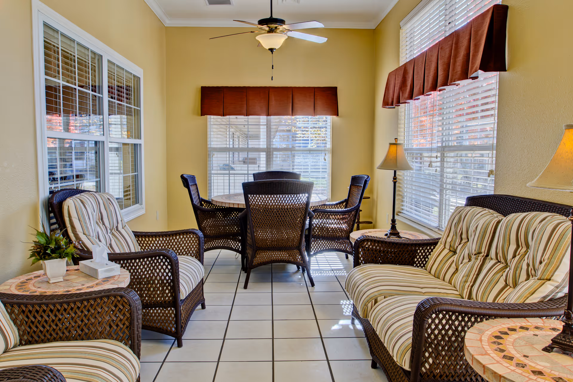 A sunlit sitting area with wicker furniture including a cushioned loveseat, two armchairs, and a round table with four wicker chairs. The room has large windows with white blinds and brown valances, beige walls, tiled floor, two side tables with lamps, and a ceiling fan with a light fixture.