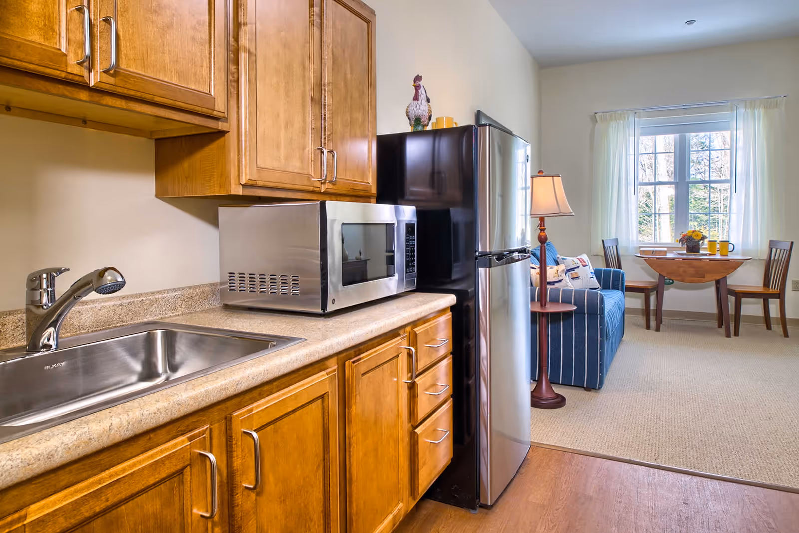 Small kitchenette with wooden cabinets, stainless steel sink, microwave and refrigerator opening into a living area with a striped sofa and a small dining table by a window.