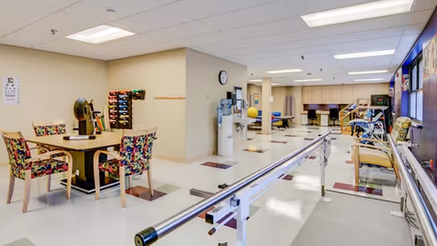 Interior of a senior living facility's rehabilitation gym showing parallel bars, seating, and exercise equipment along a long hallway.