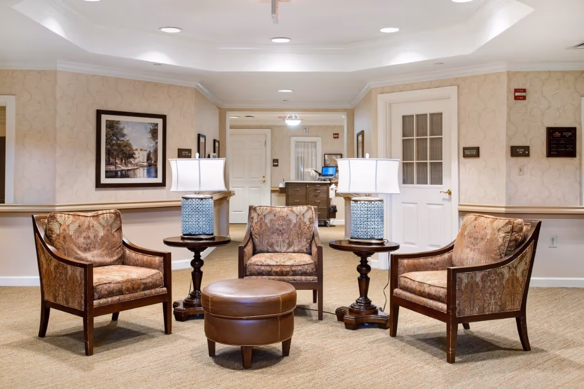 A furnished seating area with four upholstered chairs, two side tables with lamps, and a round ottoman in a common room.