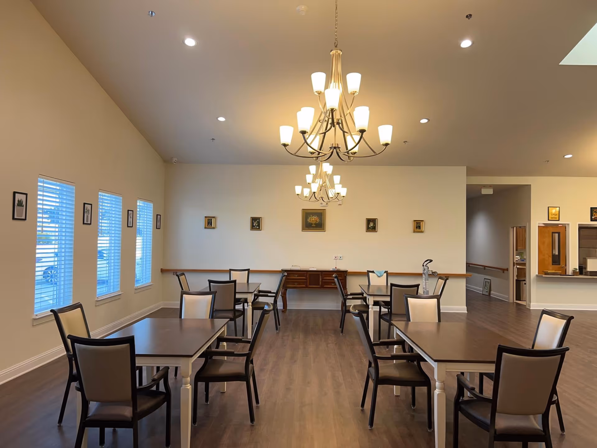 A spacious dining room with multiple rectangular tables and chairs arranged neatly. The room has light-colored walls adorned with small framed pictures and three large chandeliers hanging from the ceiling. There are several windows with blinds on the left side, and a counter area is visible in the back right corner.