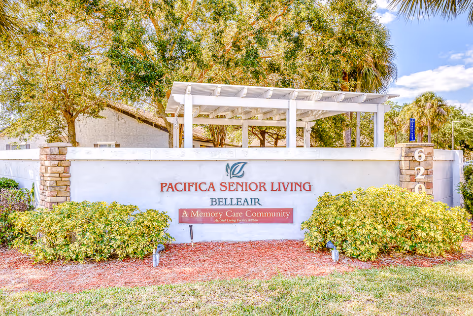 Outdoor entrance sign for Pacifica Senior Living Belleair, a memory care community, surrounded by greenery and landscaping with a white pergola structure behind the sign.