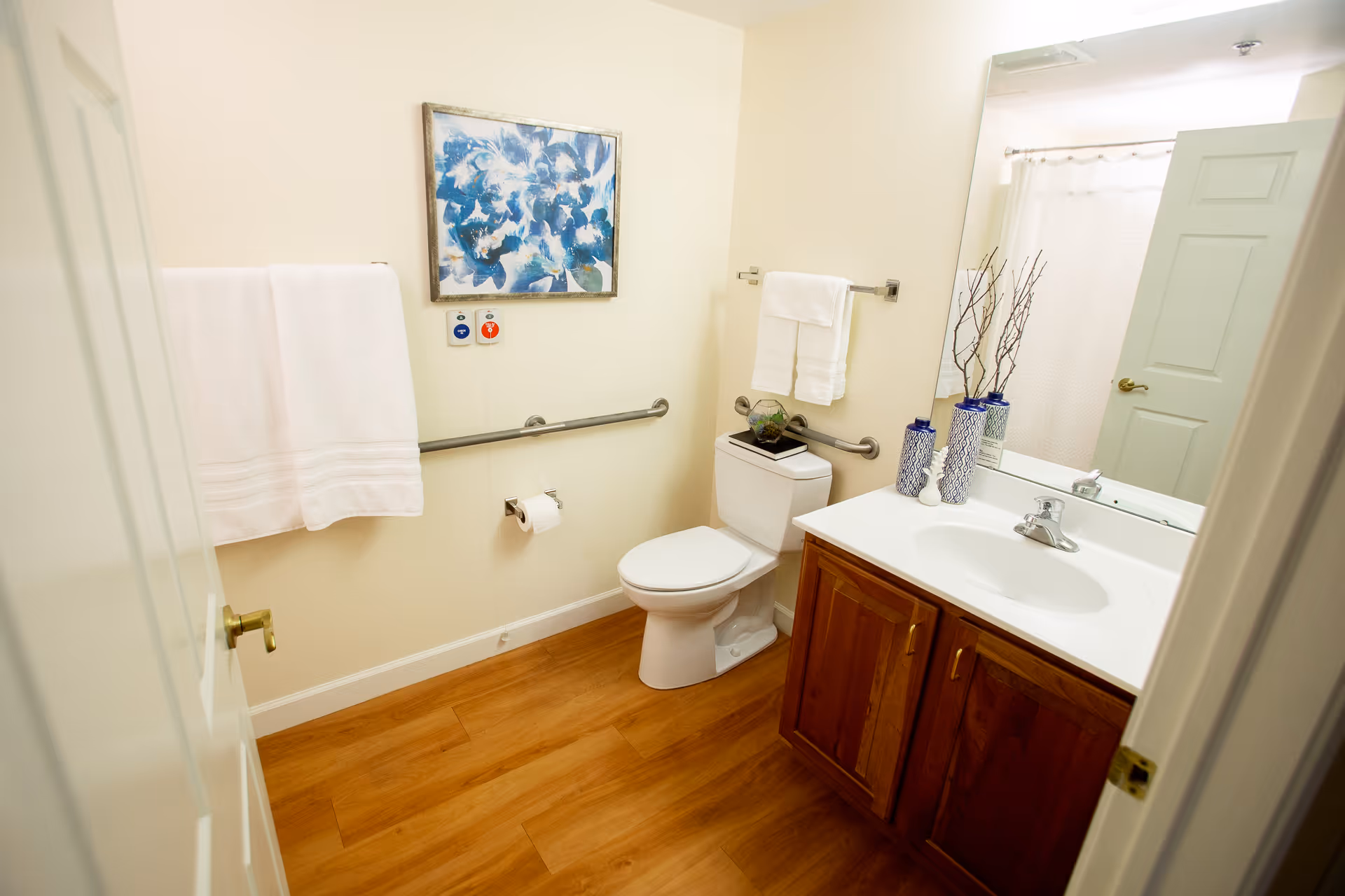 A clean bathroom with wooden flooring, a white toilet with a decorative item on top, a white sink with wooden cabinetry, a large mirror above the sink, white towels hanging on metal bars, and a blue and white floral painting on the wall.