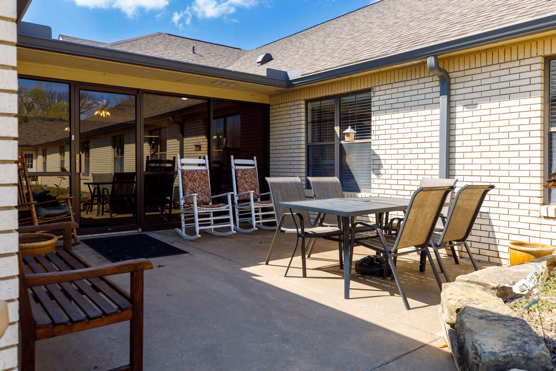 Outdoor patio area at Asbury Village with a table and four chairs, two white rocking chairs with patterned cushions, a wooden bench, and a stone planter. The patio is adjacent to a building with white brick walls and large glass doors.