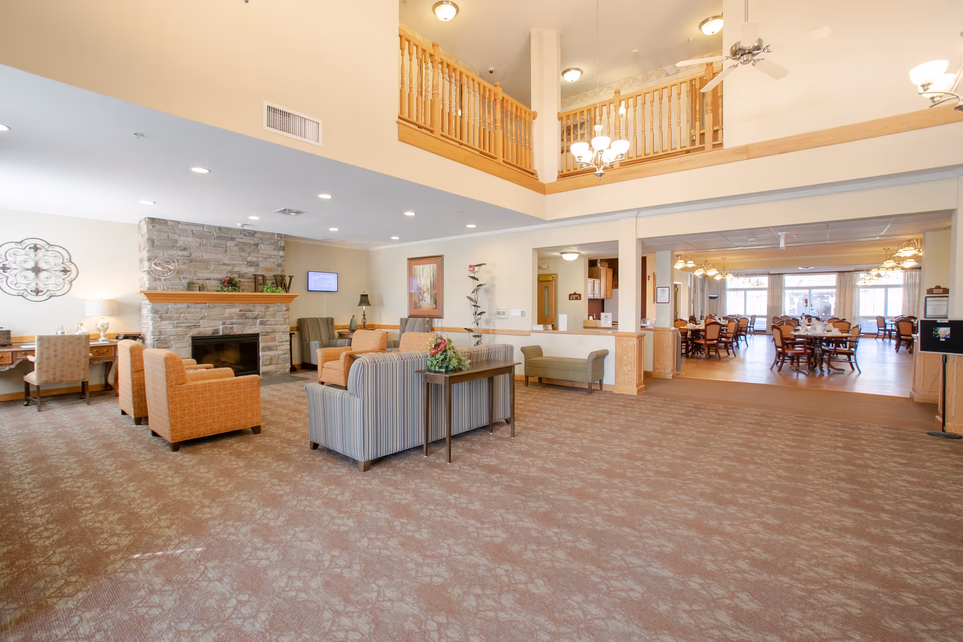 Spacious senior living facility common area with a high ceiling and wooden railing on the upper level. The room features a stone fireplace, comfortable armchairs and sofas arranged around it, a desk with a chair, and a dining area with multiple tables and chairs visible in the background. The space is well-lit with ceiling lights and chandeliers.