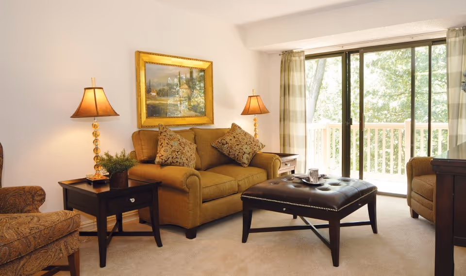 A cozy living room with a brown sofa adorned with patterned cushions, two matching table lamps on dark wooden side tables, a large framed painting on the wall, a patterned armchair, and a tufted leather ottoman in the center. The room has beige carpeting and large sliding glass doors that open to a balcony with a view of green trees outside.