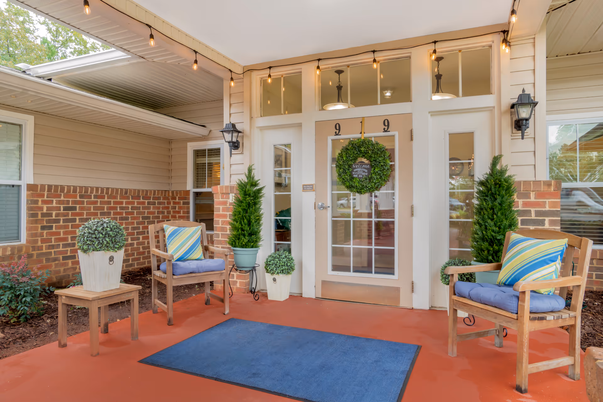 Covered entrance area of a building with a glass door decorated with a green wreath. Two wooden chairs with blue cushions and striped pillows are placed on either side of the door, along with potted plants. The floor is red with a blue mat in front of the door. String lights hang from the ceiling above.