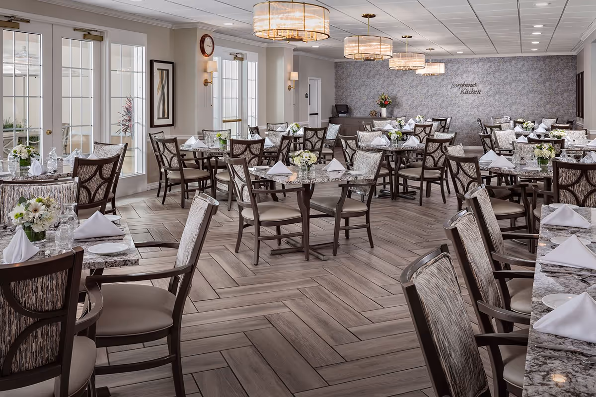A spacious and elegant dining room with multiple tables set with white napkins, plates, glasses, and floral centerpieces. The room features large windows and glass doors allowing natural light, modern chandeliers, and a wall with the text 'Josephine's Kitchen'.