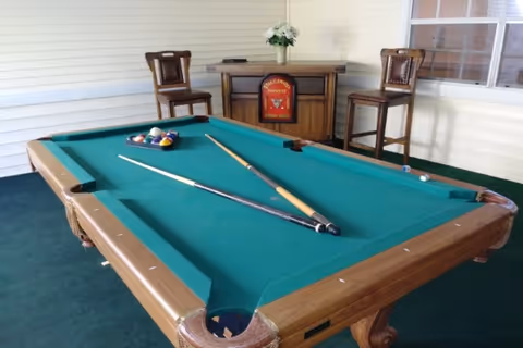 A green-felt pool table with two cues and racked balls in a covered game area with two bar stools and a small cabinet with flowers.