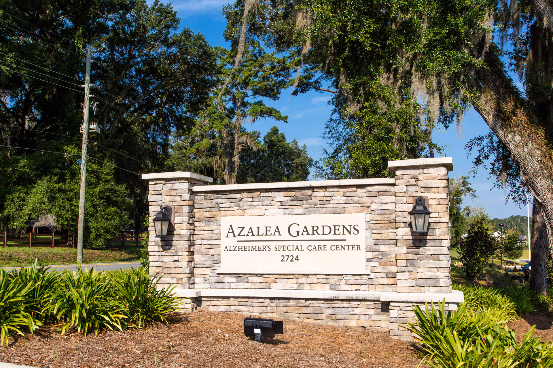 Stone sign for Azalea Gardens Alzheimer's Special Care Center with the address 2724, surrounded by greenery and trees under a clear blue sky.
