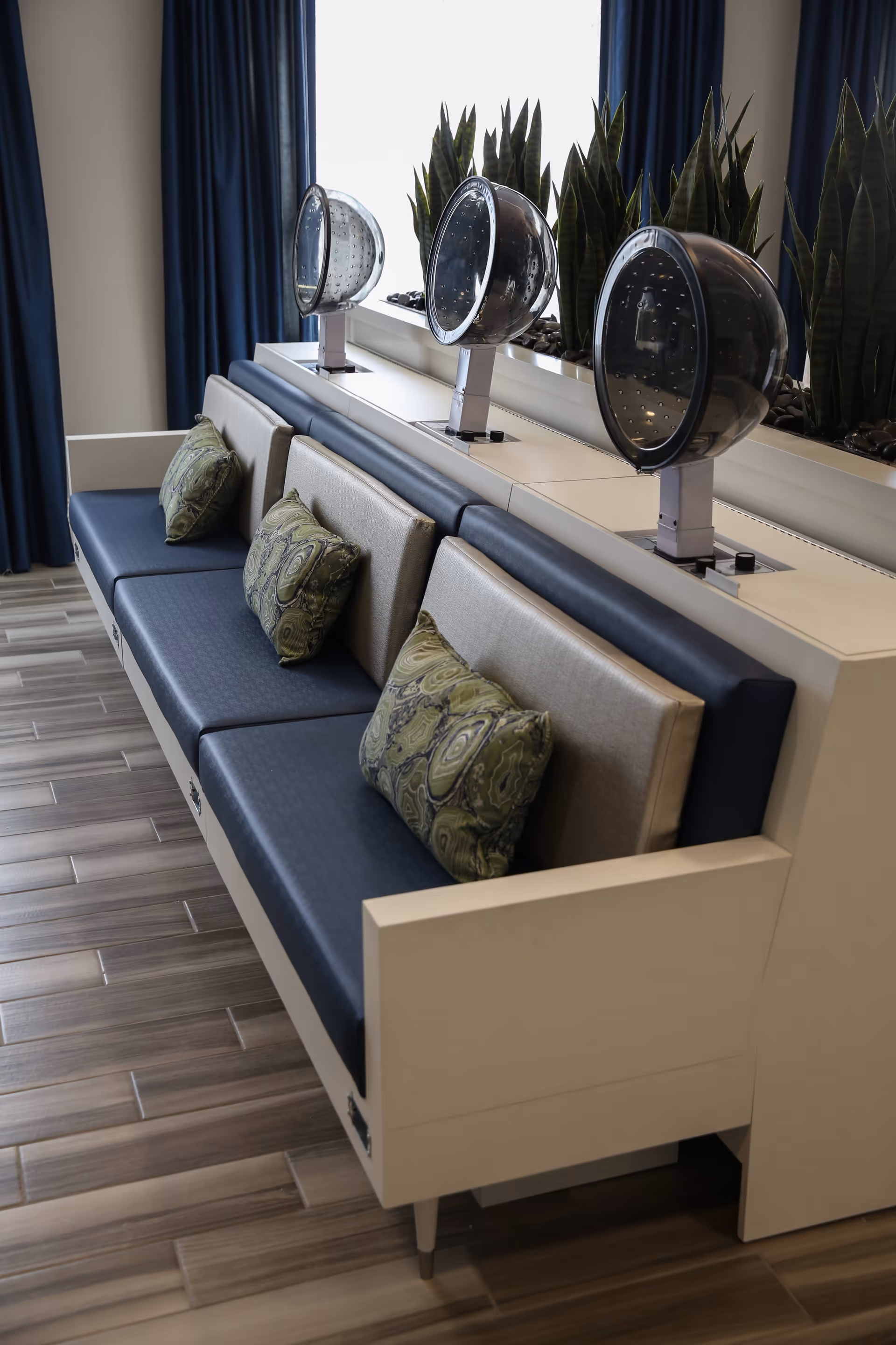 A row of three vintage-style hair drying stations with black dome hair dryers mounted on a white counter behind a long bench seat with blue cushions and decorative green pillows. Tall green plants are placed behind the dryers, and dark blue curtains frame a large window in the background.
