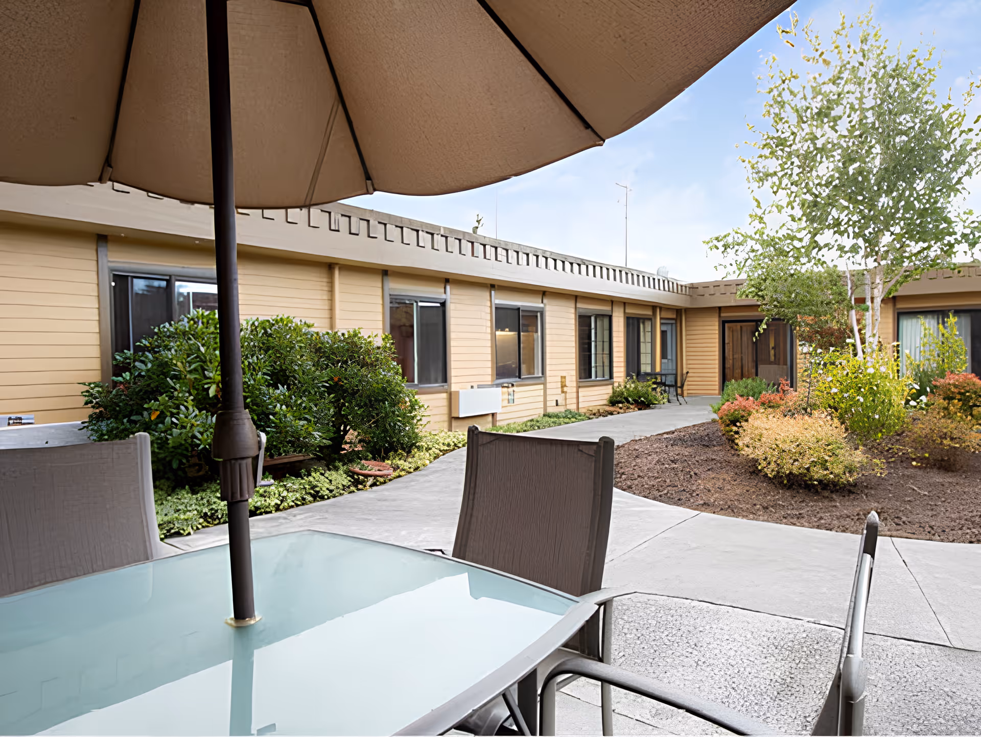 Outdoor courtyard area with a glass table and chairs under a large umbrella, surrounded by a garden with bushes and trees, and a beige building with multiple windows in the background.
