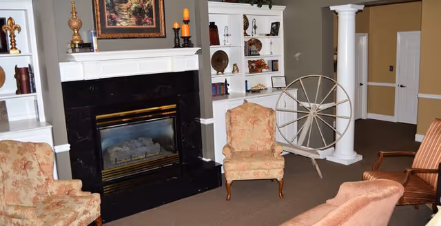 A cozy senior living common area featuring a black marble fireplace with a white mantel, flanked by built-in white bookshelves displaying decorative items. There are several upholstered armchairs with floral and striped patterns arranged around the fireplace. The room has beige walls, a brown carpet, and a large decorative spinning wheel near a white column.