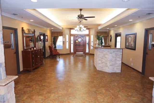 A spacious and well-lit reception area with a wooden front door at the far end, a chandelier and ceiling fan overhead, a stone-faced reception desk on the right, a wooden dresser with a mirror on the left, and a red upholstered chair near the door. The walls are painted beige with framed artwork and windows with curtains.