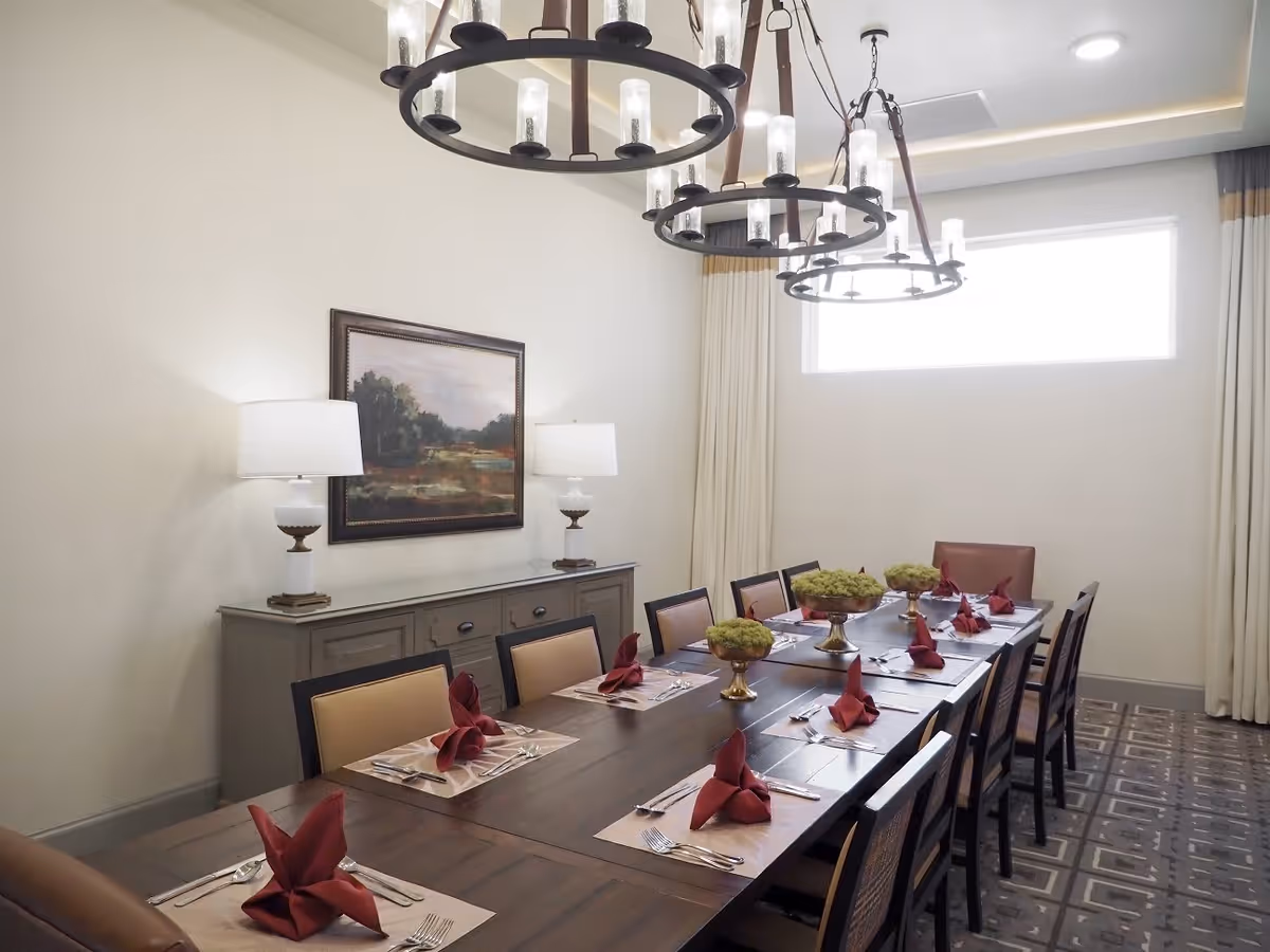 A formal dining room with a long wooden table set for twelve people. Each place setting has a beige placemat, silverware, and a folded red napkin. Three decorative centerpieces with green moss are placed along the middle of the table. The room features two large circular chandeliers hanging from the ceiling, a sideboard with two white lamps, and a framed landscape painting on the wall. A large window with white curtains allows natural light into the room.